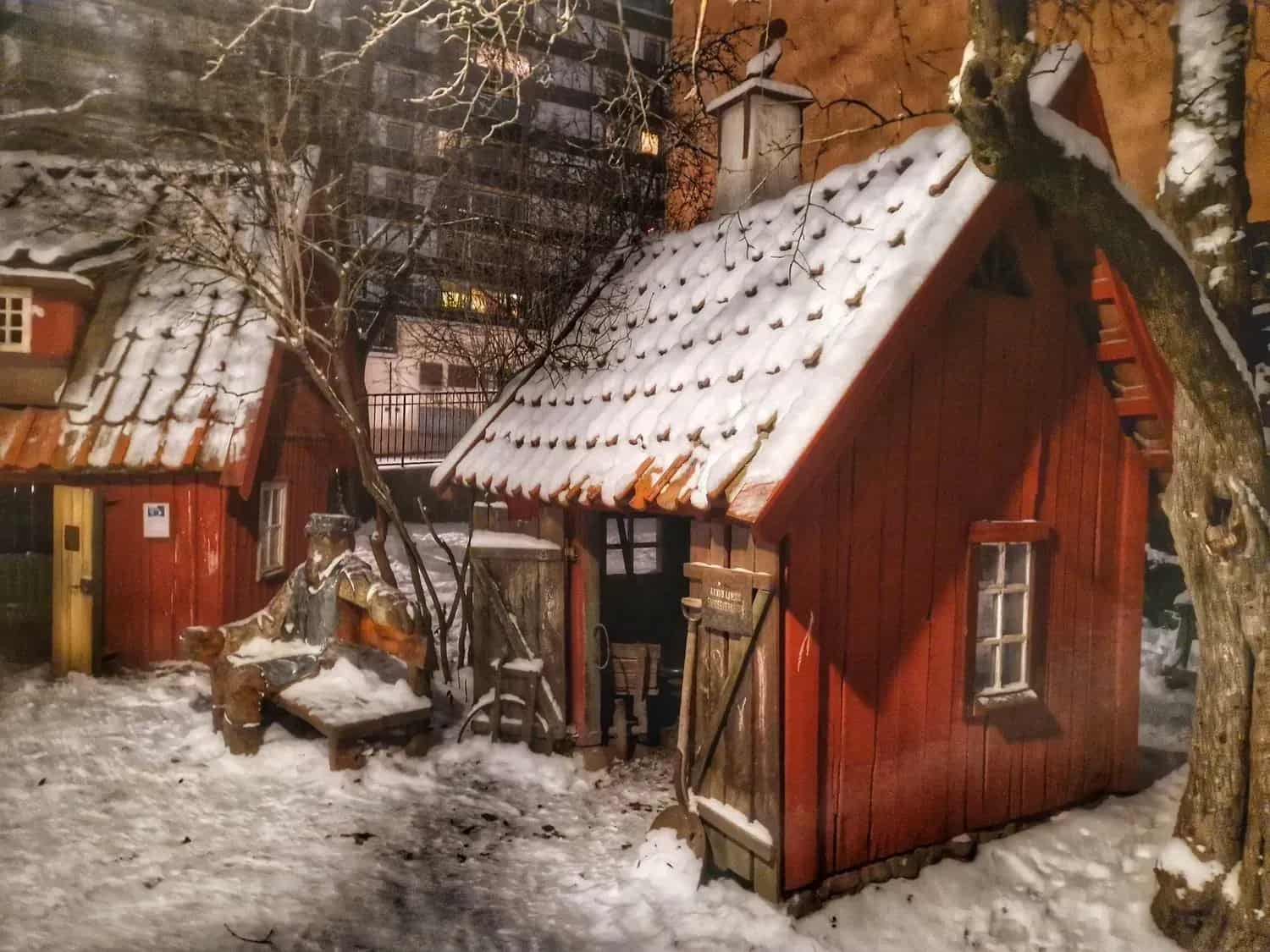 Två röda stugor med snö på taken. Snö på marken. Träd och hus i bakgrunden