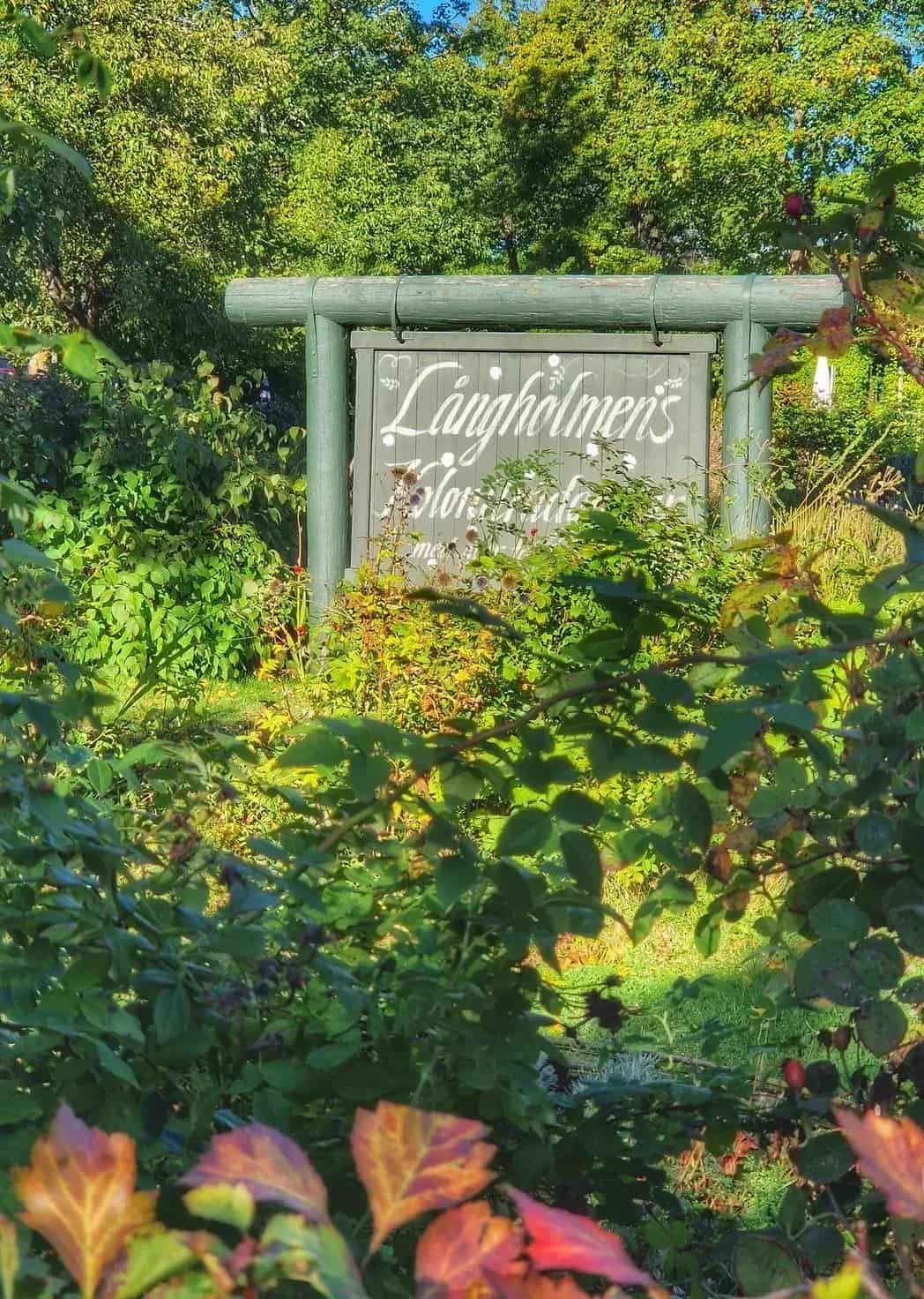 Träskylt med texten "Långholmens floristträdgård" bland grönska