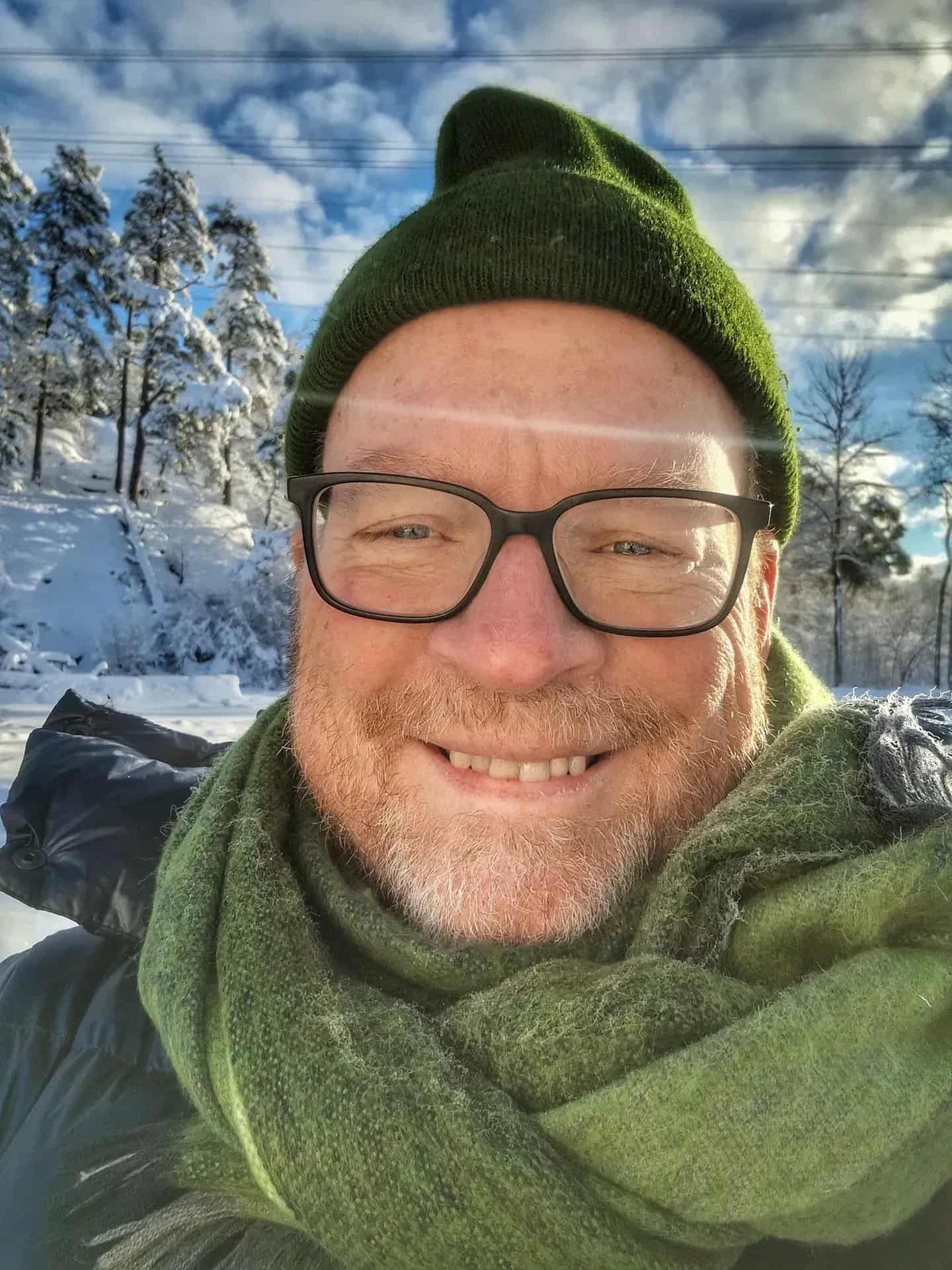 Smiling man with glasses, green hat and scarf outdoors. Snow and trees in the background