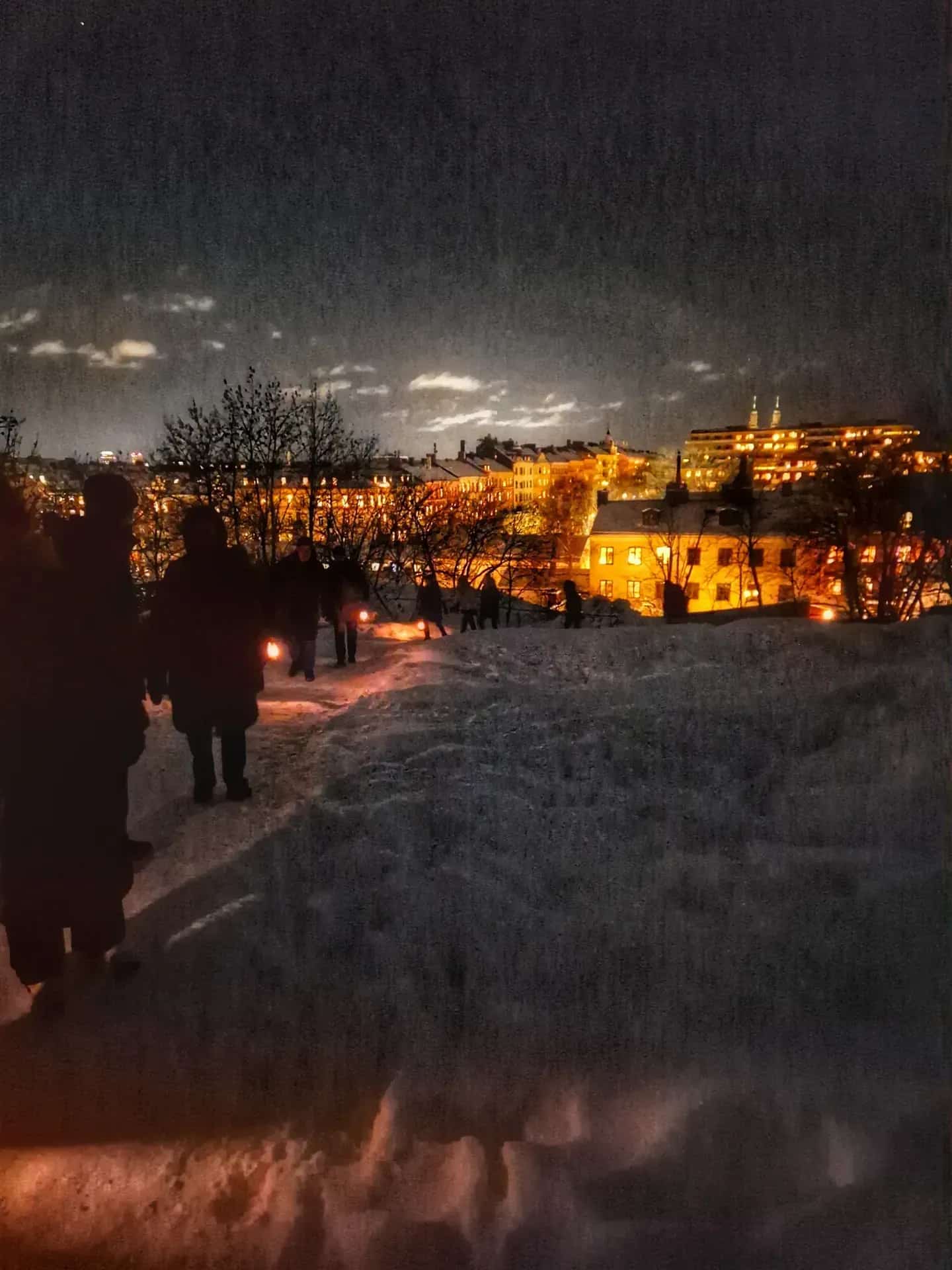 Mörkt vinterlandskap. Silhuetter av människor med lyktor i snön. Upplysta byggnader i bakgrunden