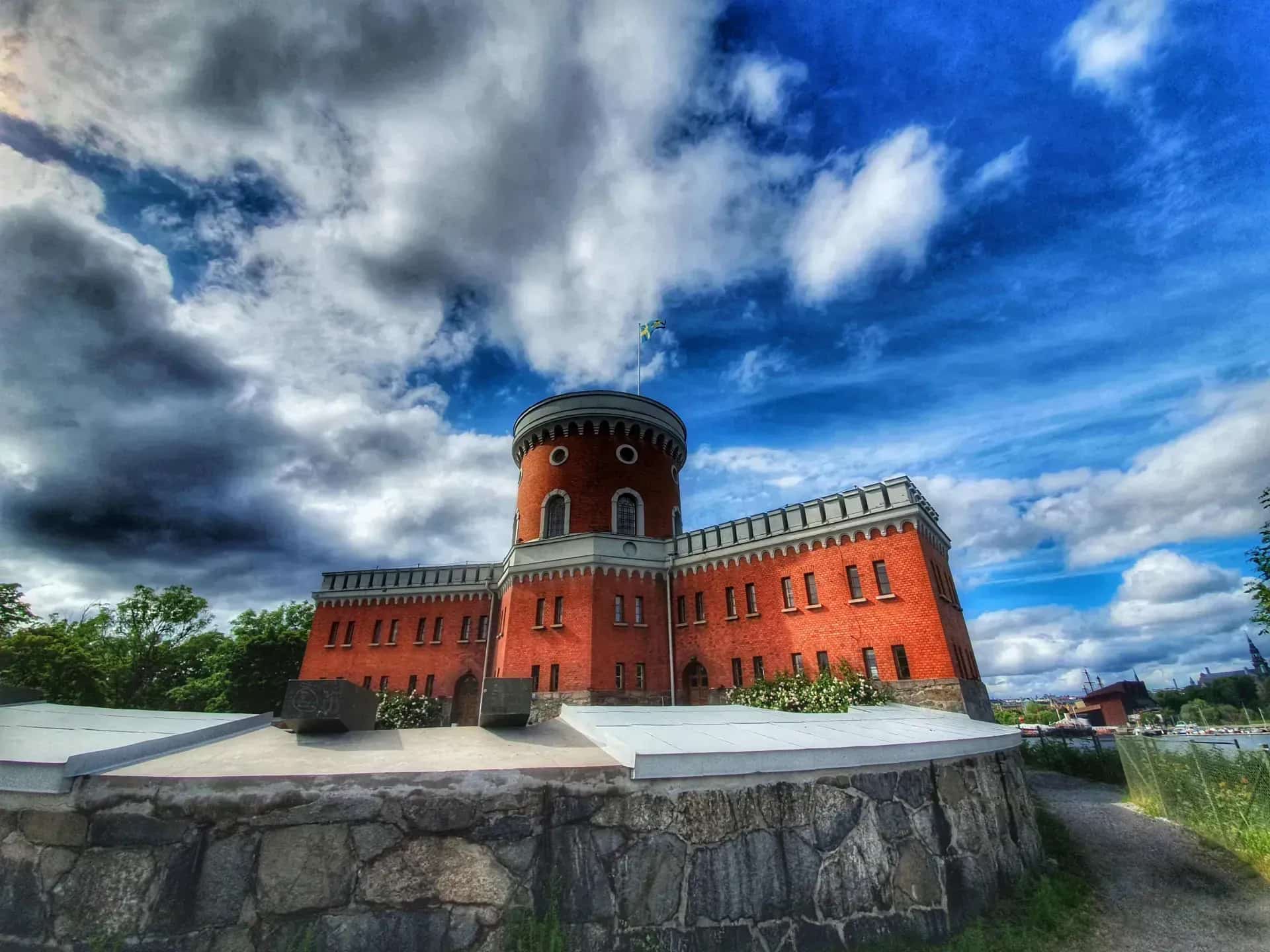 Kastellholmen, en ö i Stockholm. Rött tegelhus med torn under blå himmel med moln. Svensk flagga. Stensockel