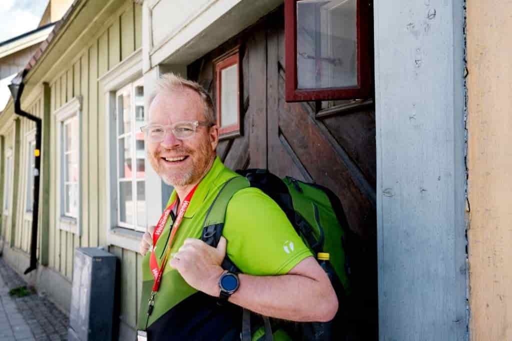 Man with backpack and glasses smiles. He stands by a wooden door
