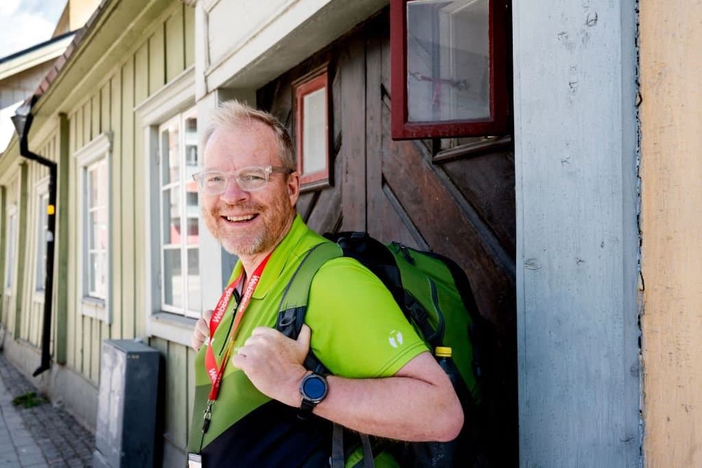 Smiling man with backpack, green sweater and glasses in front of house with windows