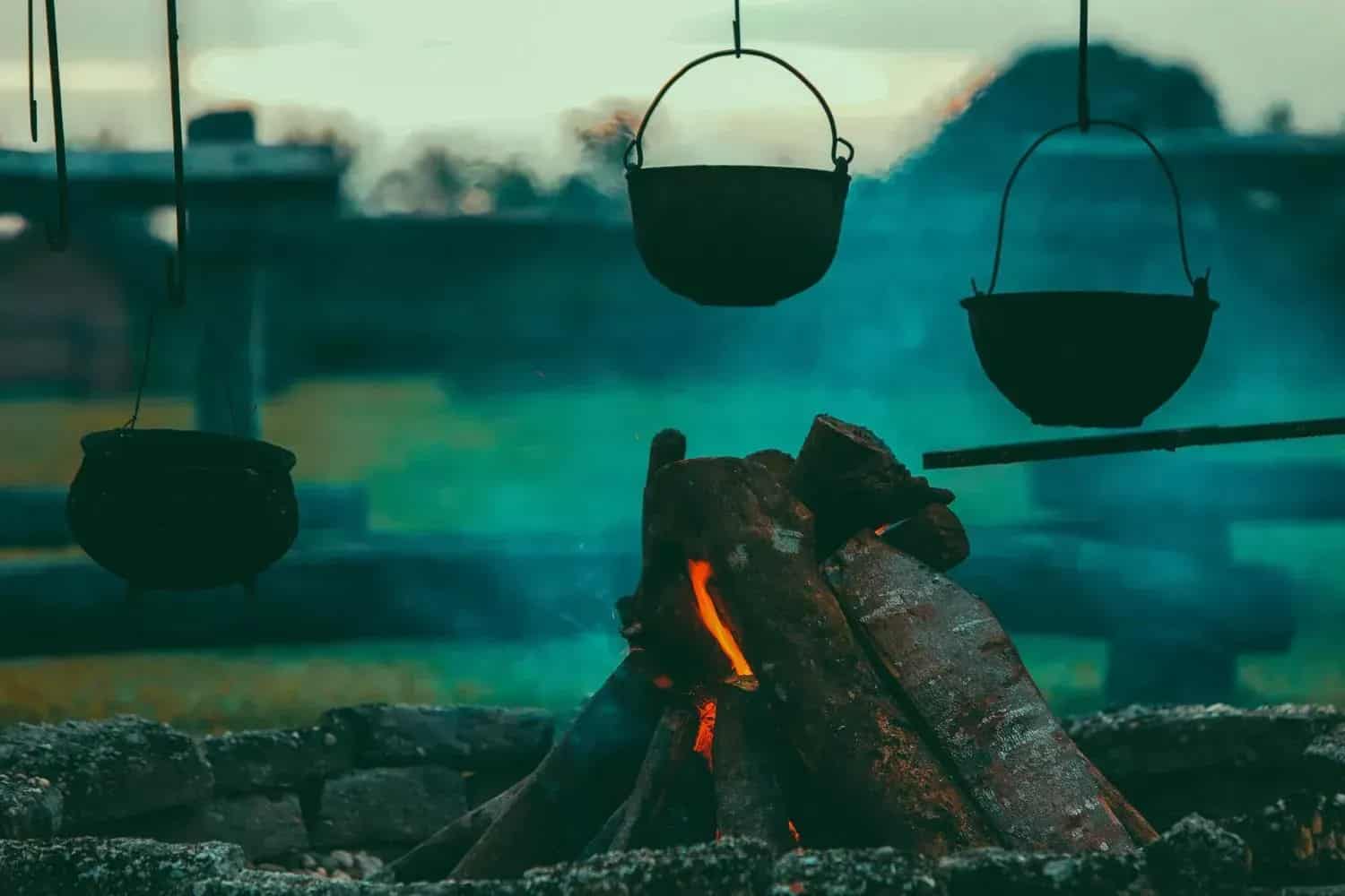 Fire with wood in stone circle. Three black pots hang above. Smoke