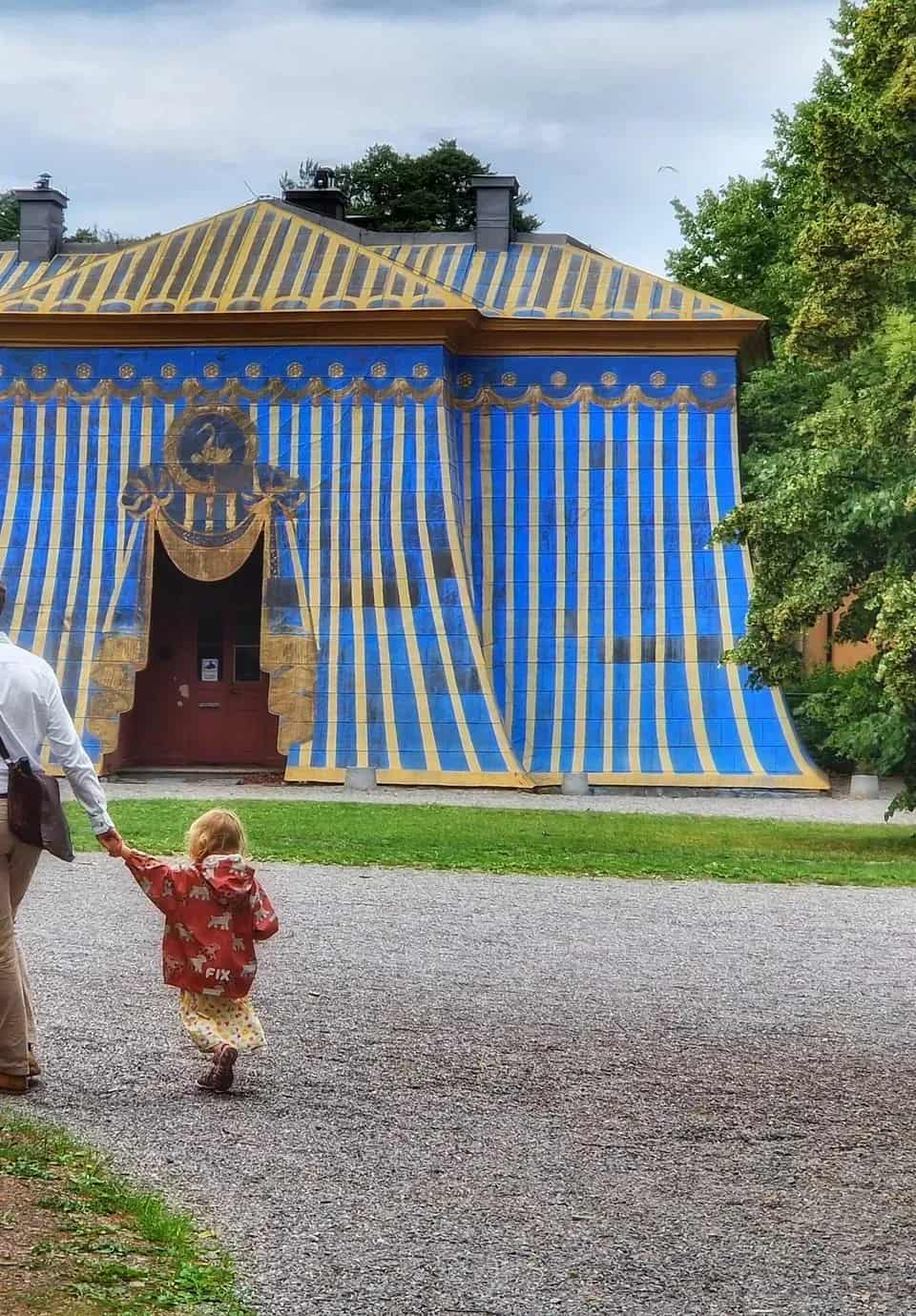 Man och barn hand i hand framför byggnad målad som tält i blått och gult