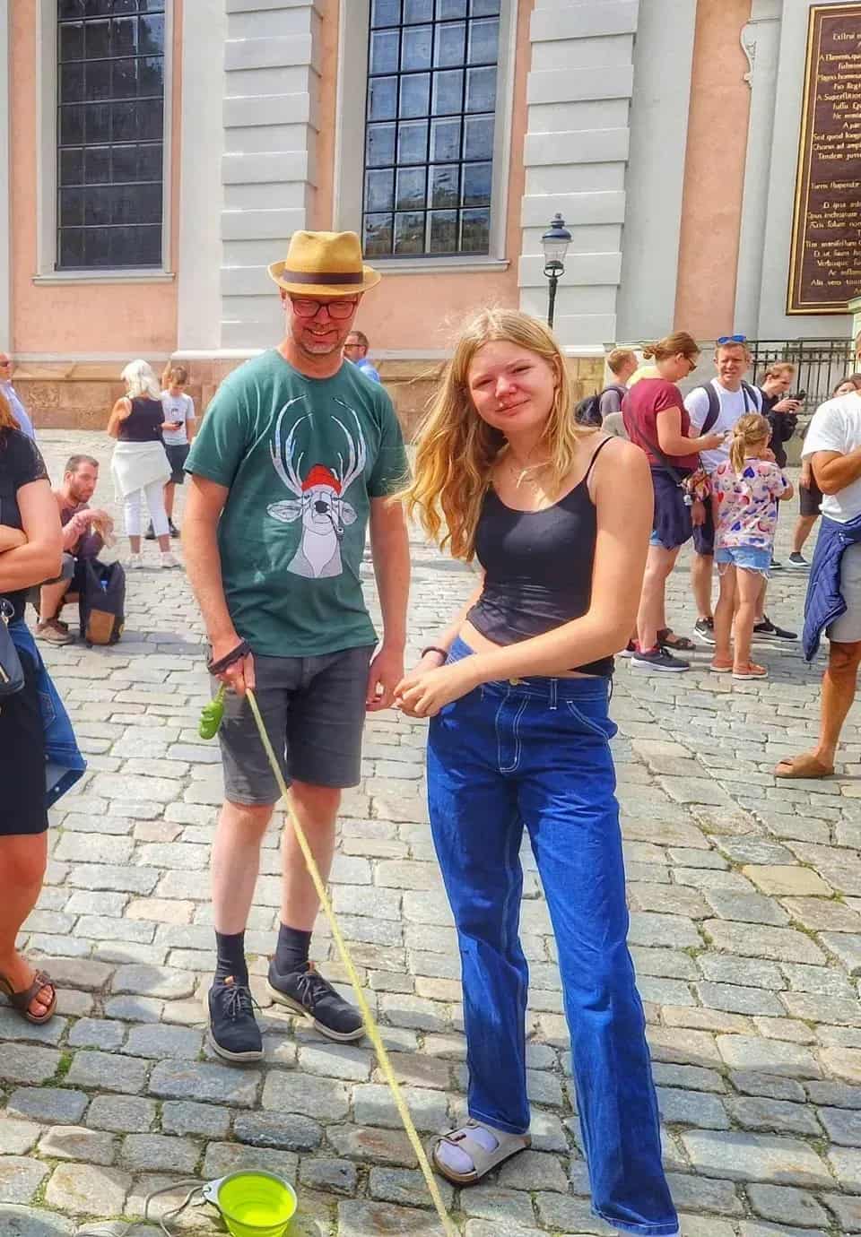 Kvinna och man på kullersten. Mannen har hatt och t-shirt med ren. Kopplad grön skål. Flera personer i bakgrunden