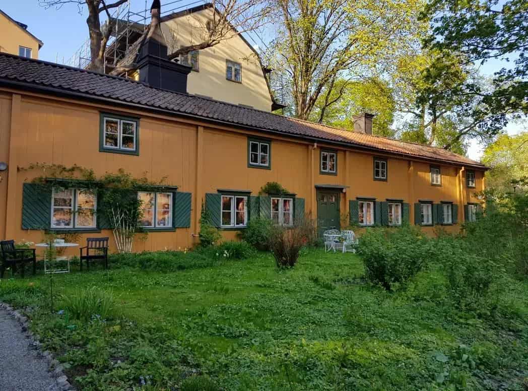 Lågt, gult hus med grön dörr och gröna fönsterluckor. Gräsbevuxen mark framför. Träd i bakgrunden