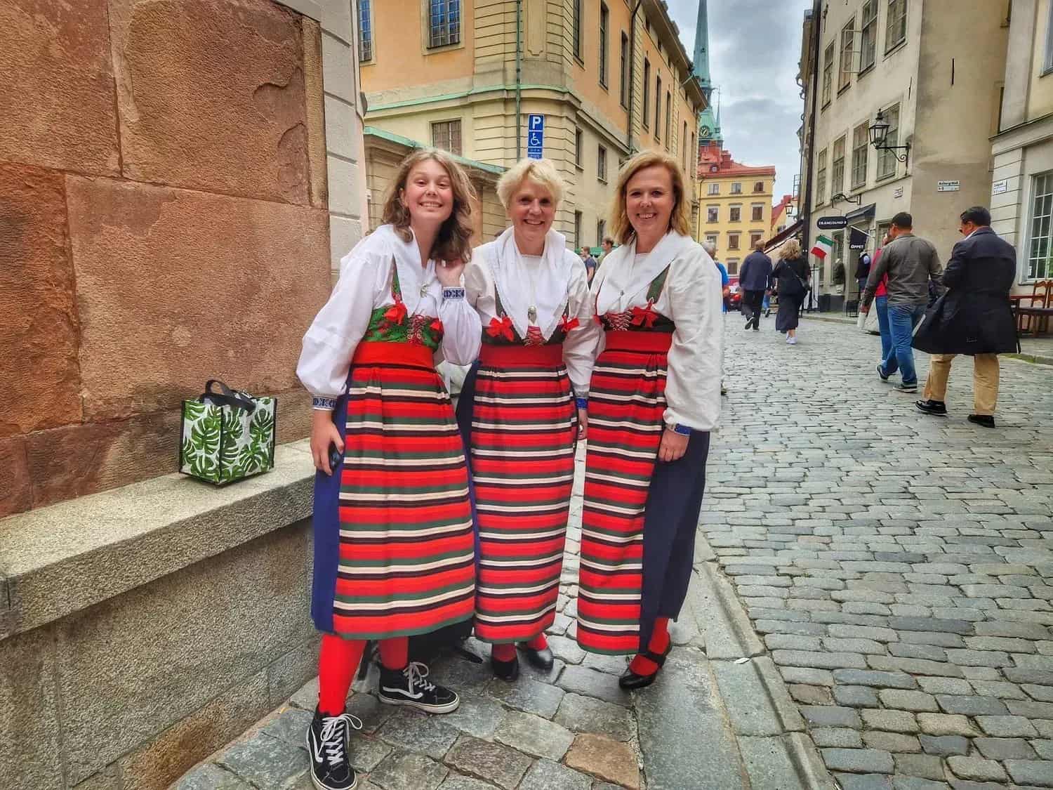 Three women in folk costume stand on a cobblestone street. Houses in the background
