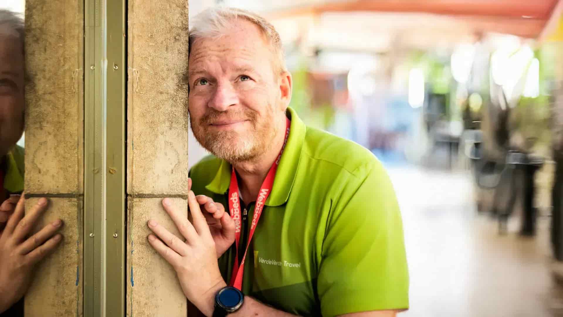 Man in green shirt with red band around his neck leans against concrete pillar