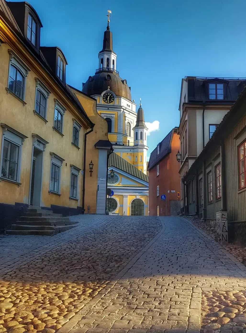 Gata med kullerstenar leder upp mot kyrka med torn och klocka. Hus på båda sidor. Blå himmel