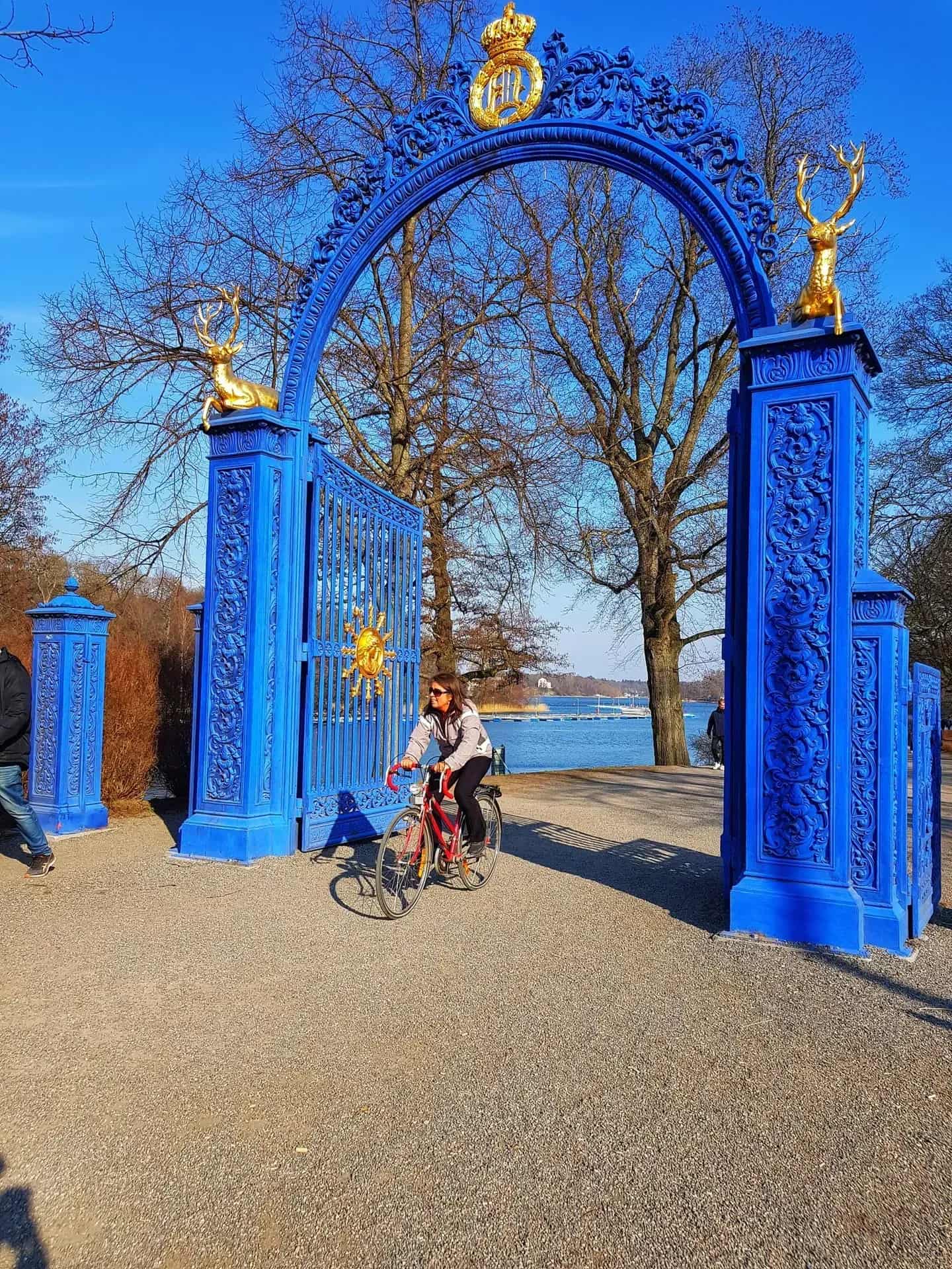 Blue gate with gold decor, sun symbol and deer. Woman cycling through. Water in the background