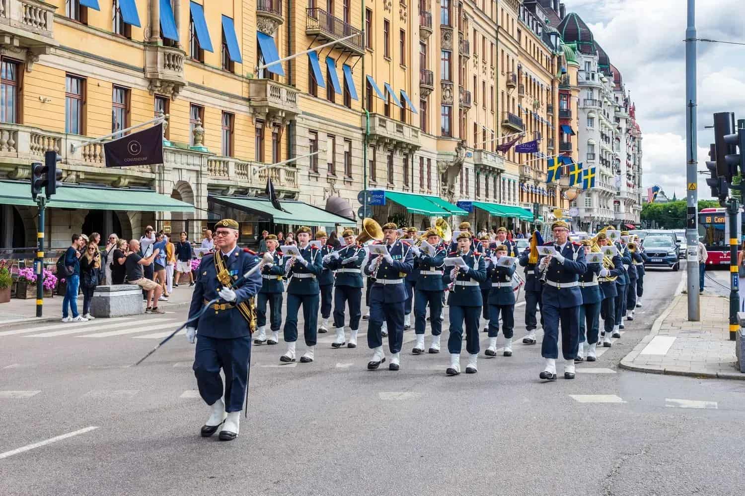 Musikkår i uniform marscherar på gata. Byggnader i bakgrunden