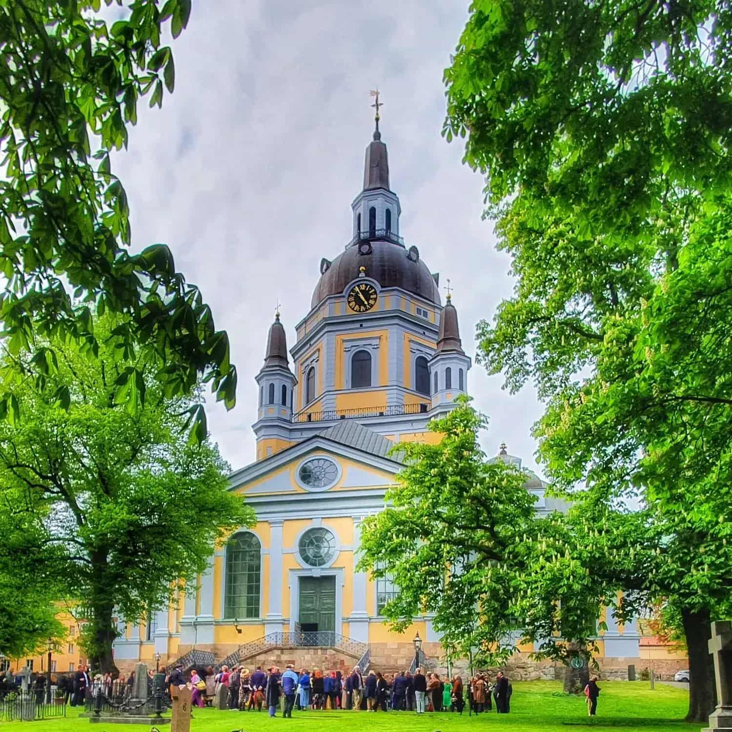 Church with tower and dome, surrounded by trees and people. Lawn and gravestones in the foreground